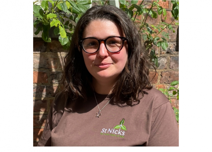 Anna is smiling into the camera, she is a white lady with brown hair that is semi curly/wavy and shoulder length. She is wearing round, dark rimmed glasses and a butterfly necklace over her t shirt. The tshirt is brown with the st nicks logo.