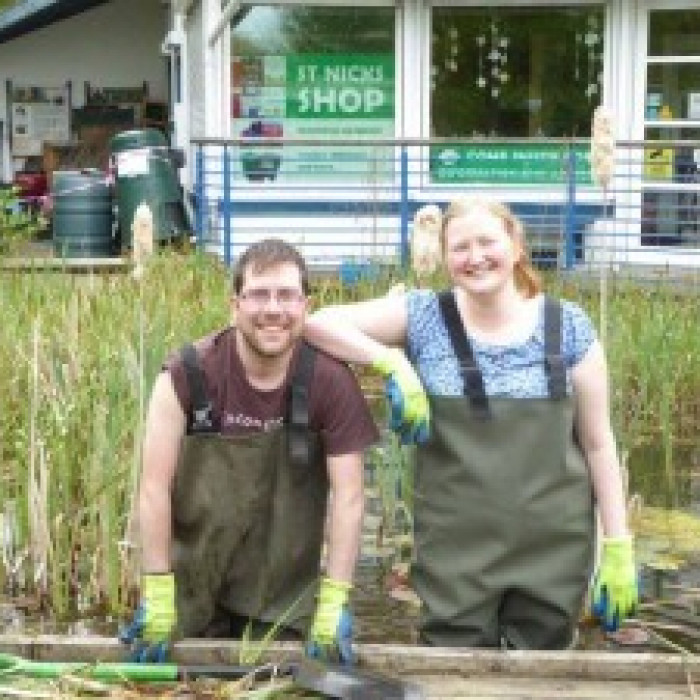 Park Rangers Nick and Harriet both wearing waders and gloves stand in the pond and smiling. Nick is on the left and leaning forwards holding the edge of our pond dipping platform, he is a young, white male with stubble and glasses. Harriet has her elbow o