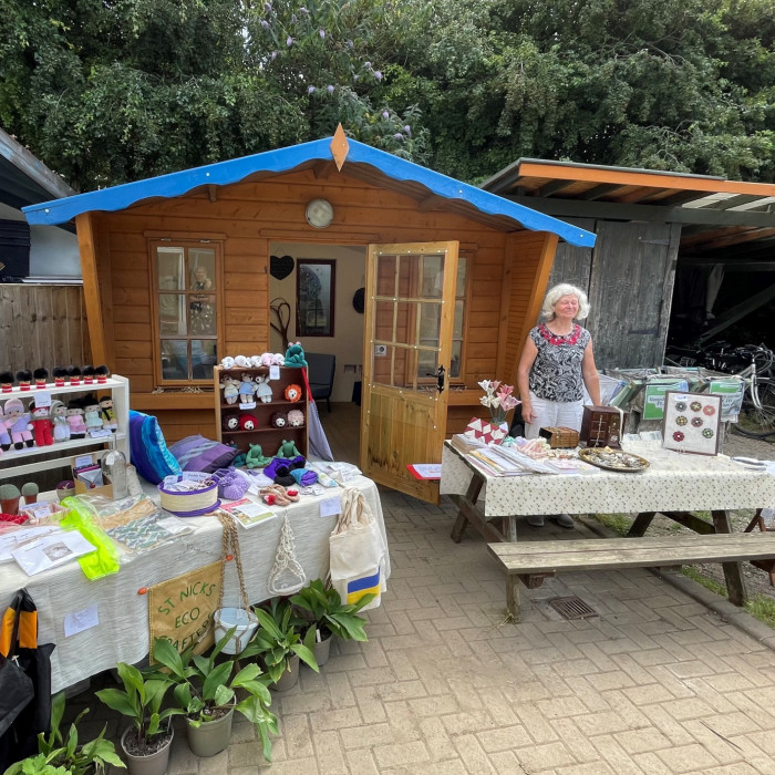 Two stalls sit outside the eco-chalet. They are full of handcrafted, upcycled items for sale from teh eco-crafters. An eco-crafter stands smiling behind the stall.