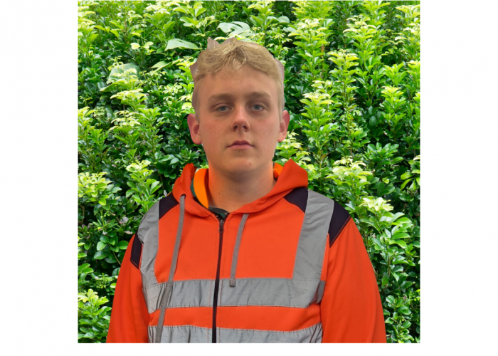 A young male with blonde hair looks straight into the camera, he is wearing orange hi vis and stands in front of a bush