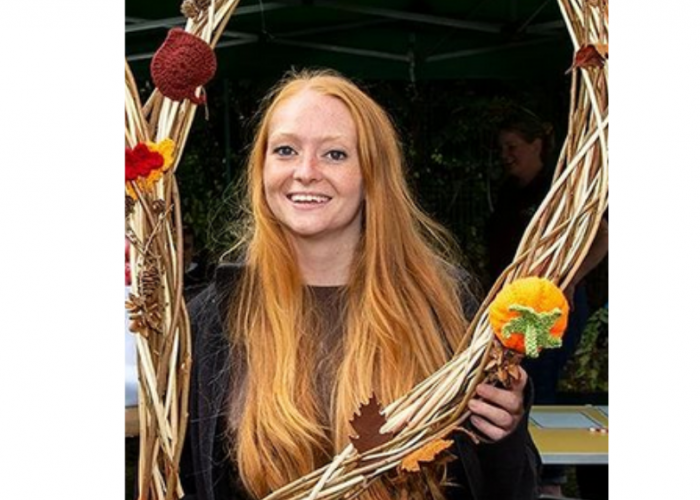 Maria Gill is a young red haired freckled woman. Her hair is incredibly long and straight. She is wearing eyeliner and smiling at the camera. She is holding a wicker heart that has woolen pumpkins and leaves sewed onto it.