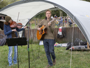 A man and woman stand under a gazebo. The man with blonde hair is looking into the camera and playing a guitar. The woman is stood behind a sheet music stand and looking off camera as she plays a violin.