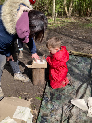 a little boy with light brown hair and white skin dressed in a red overall is kneeling holding a hammer. A female adult is stood holding some cloth in place. They are outside in the woodland