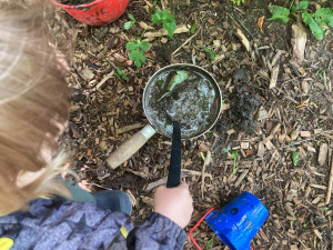 a view from above a child who is stirring a metal pot with a plastic ladle. The pot is filled with water and mud