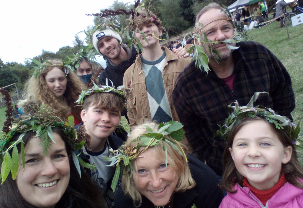 A mix of men, women and children smile into the camera which is held selfie style. They are all wearing floral crowns made from foraged materials such as leaves and flowers.