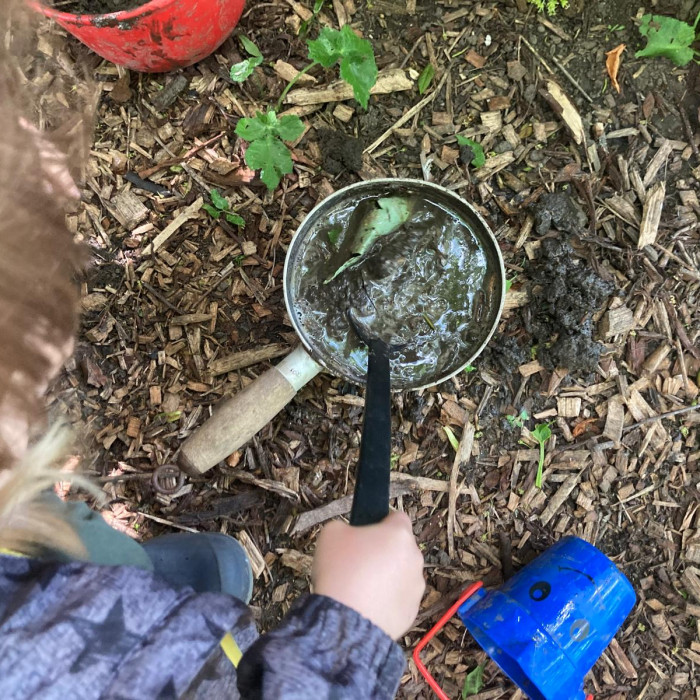 a view from above a child who is stirring a metal pot with a plastic ladle. The pot is filled with water and mud
