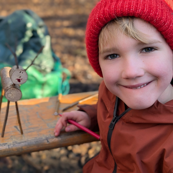 a young child with blonde fringe wearing a coat and a red knitted hat smiling sat next to a reindeer craft made with twigs, the boy is holding a felt tip pen and the reindeer has a hand drawn smiley face on it