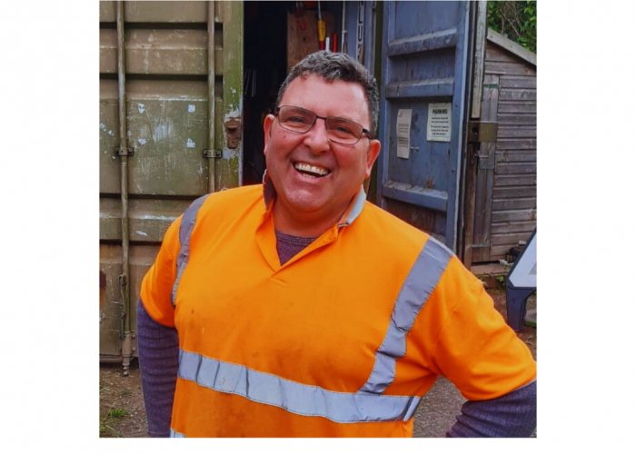 Mark Claridge, a man with a large cheeky grin, dark hair and glasses grins at the camera. He has his hands on his hips and is wearing a long sleeved blue grey tshirt under a high vis orange polo shirt.