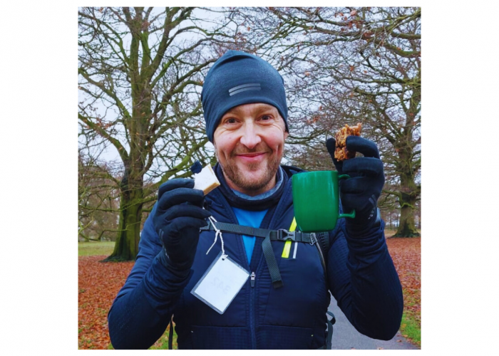 Andy, a man smiling at the camera, wearing outdoors walking gear and holding a mug and a sandwich, trees in the background