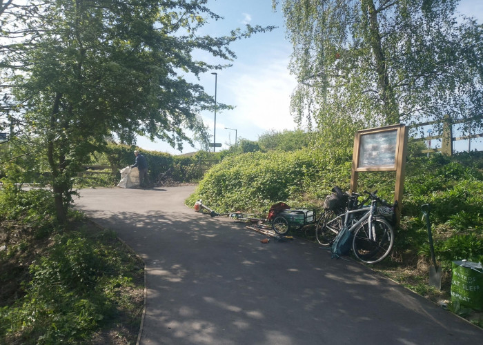 Solar System Greenway. Person in background holding large bag containing greenwaste. Cycle path lined with trees and vegetation. There is a bike and trailerleaning against a sign in a wooden frame. Tools lay alongside the edge of the path . It is a sunny