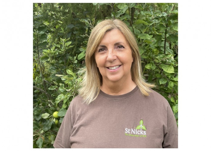 Sara has shoulder length blonde hair and brown eyes and is smiling at the camera. She is wearing a St Nicks t shirt and standing in front of an apple tree.