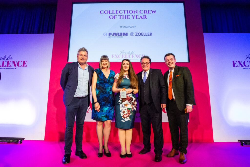 a row of smiling people look down into the camera. There are three men and two women. The men are in suits the women blue and green dresses One of the women is holding an award. The board behind them says 