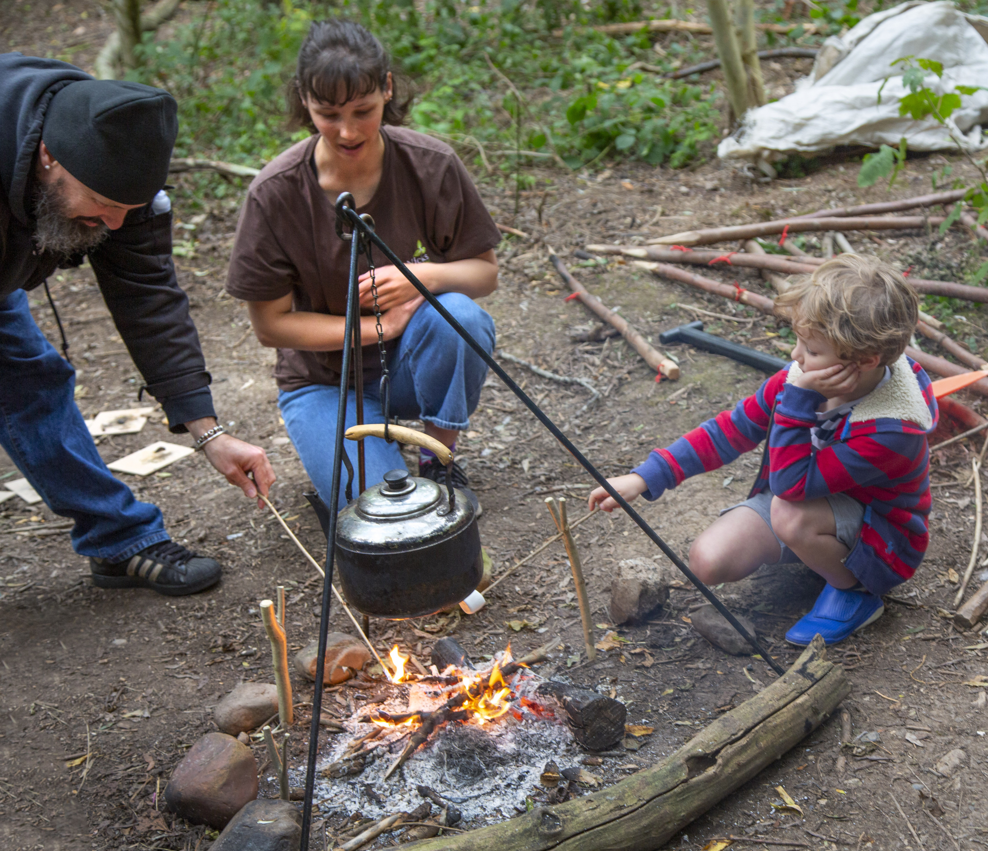 A bearded white man, a young brunette woman and a small boy crouch around n open fire over which is a black kettle suspended. Beneath the kettle and above the flames they are roasting marshallows on sticks.